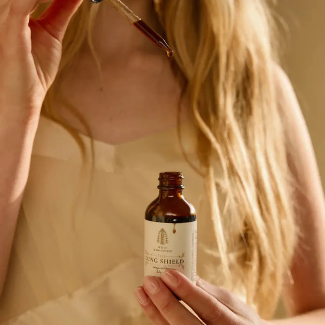 Person holding a dropper with liquid over a bottle labeled 'Lung Shield' against a blurred background.