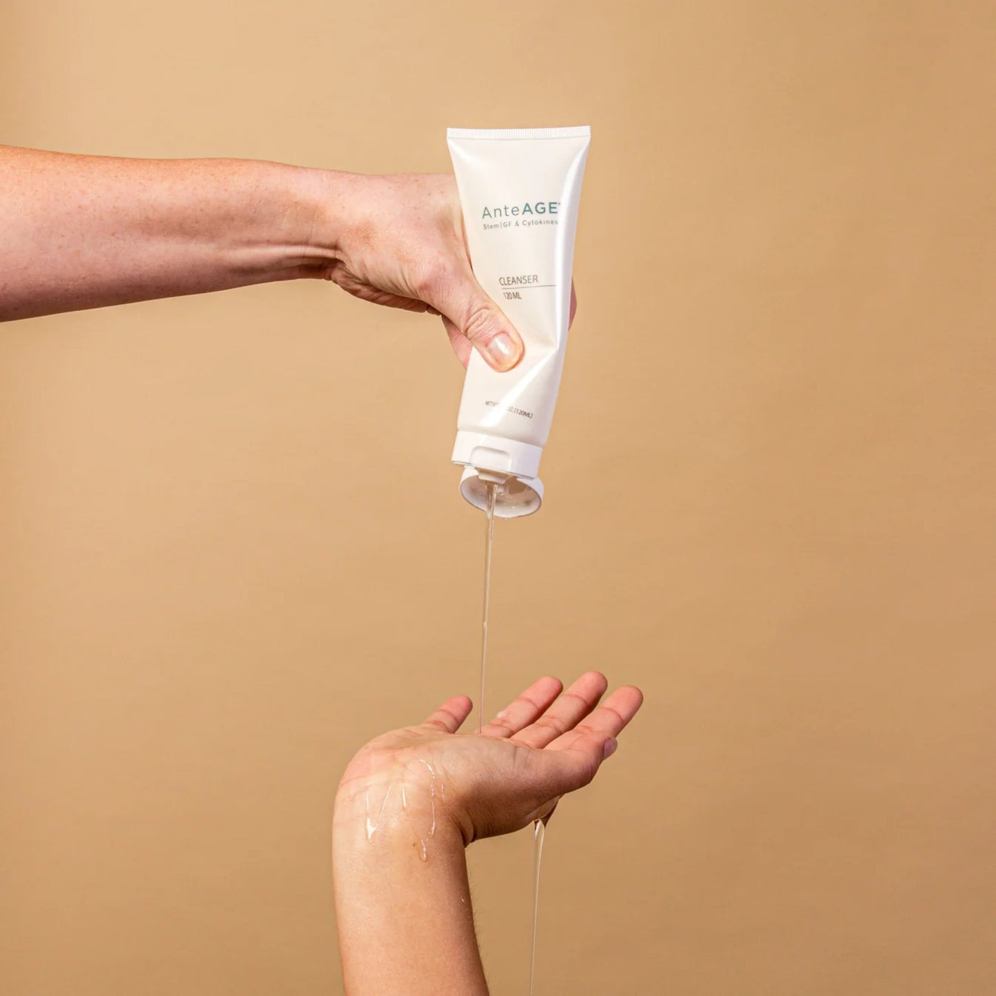Hand holding a tube of AnteAGE skincare product with cream flowing onto another hand against a beige background