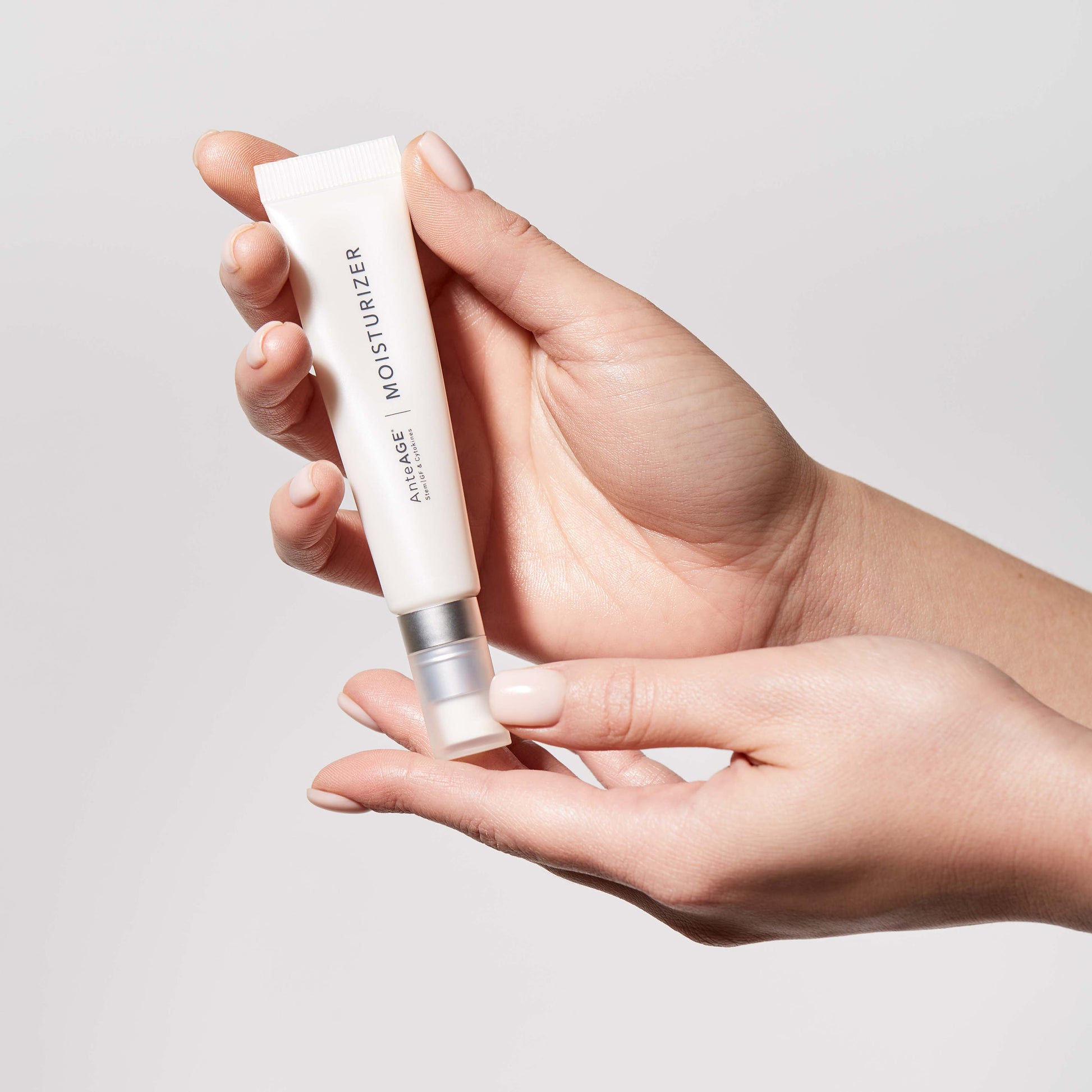 Hand holding a white Anteage moisturiser tube against a light gray background
