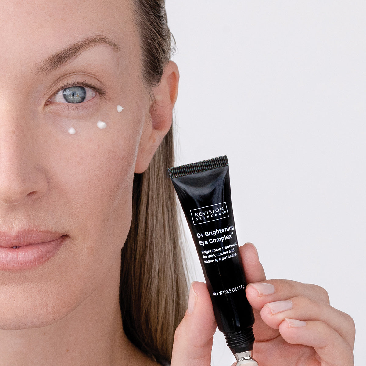 Woman applying eye cream to her face with a close-up of the Revision Skincare tube.