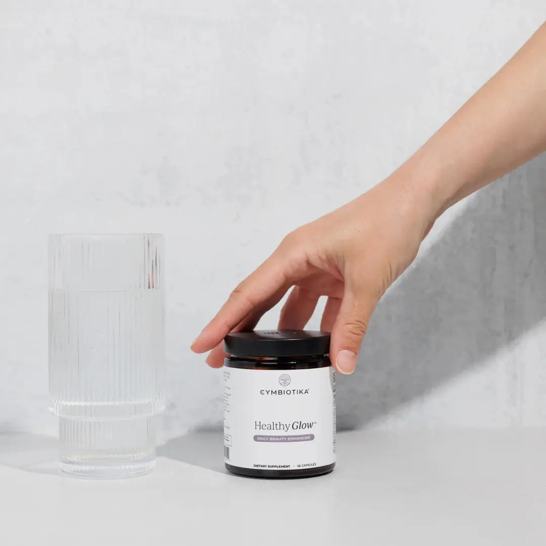 Hand holding a Cymbiotika 'Healthy Glow' supplement jar on a light gray background