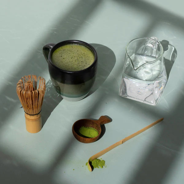 Matcha tea set with a cup, whisk, scoop, and glass of water on a light surface.