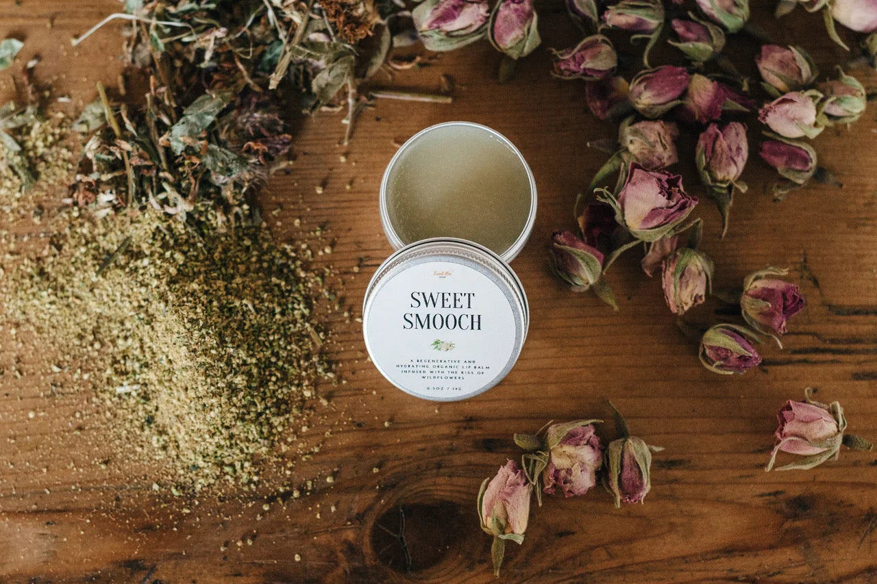 Small tin labeled 'Sweet Smooch' on a wooden surface with dried flowers and herbs.