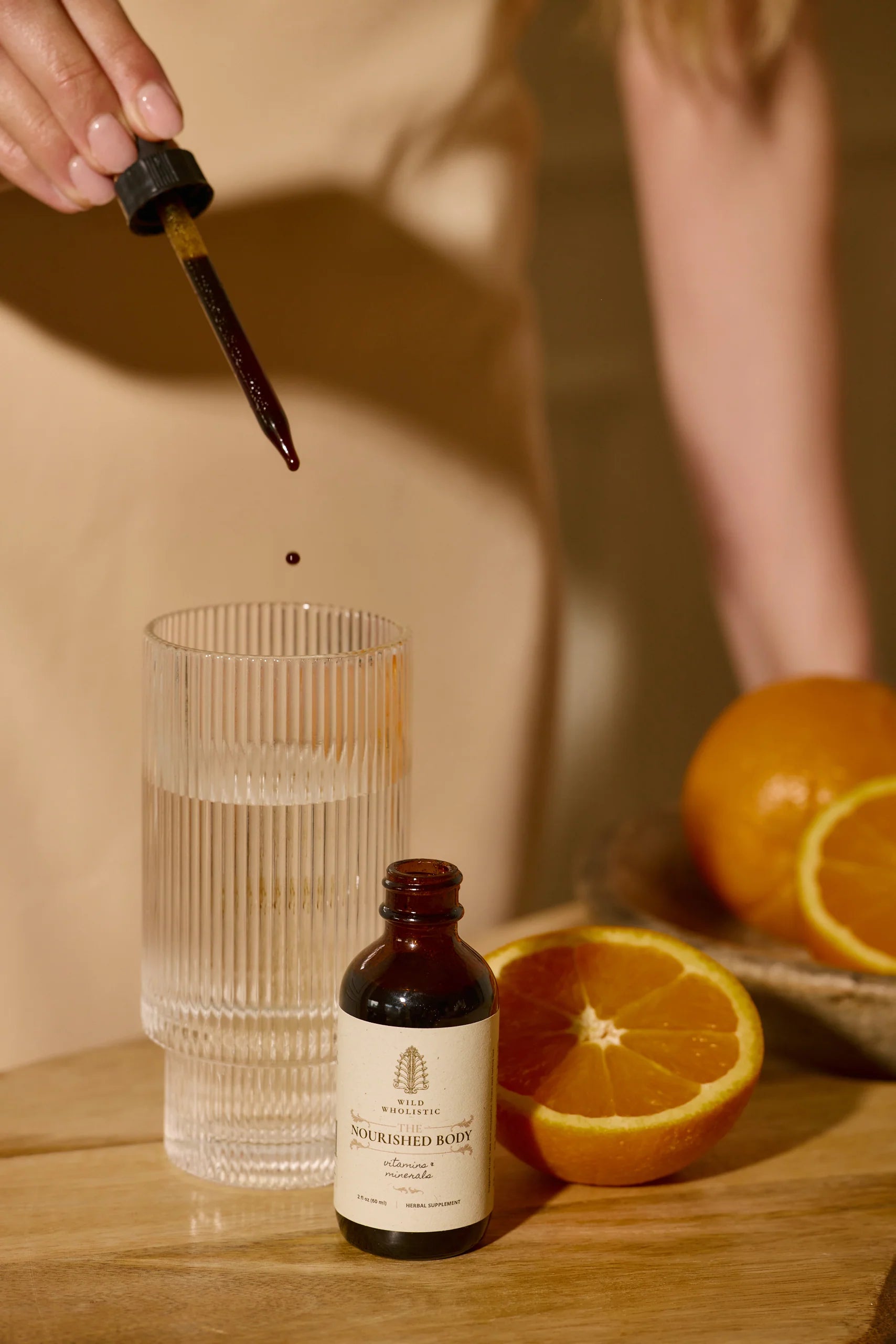 Bottle of Nourished Body oil with dropper, glass, and oranges on a wooden surface.
