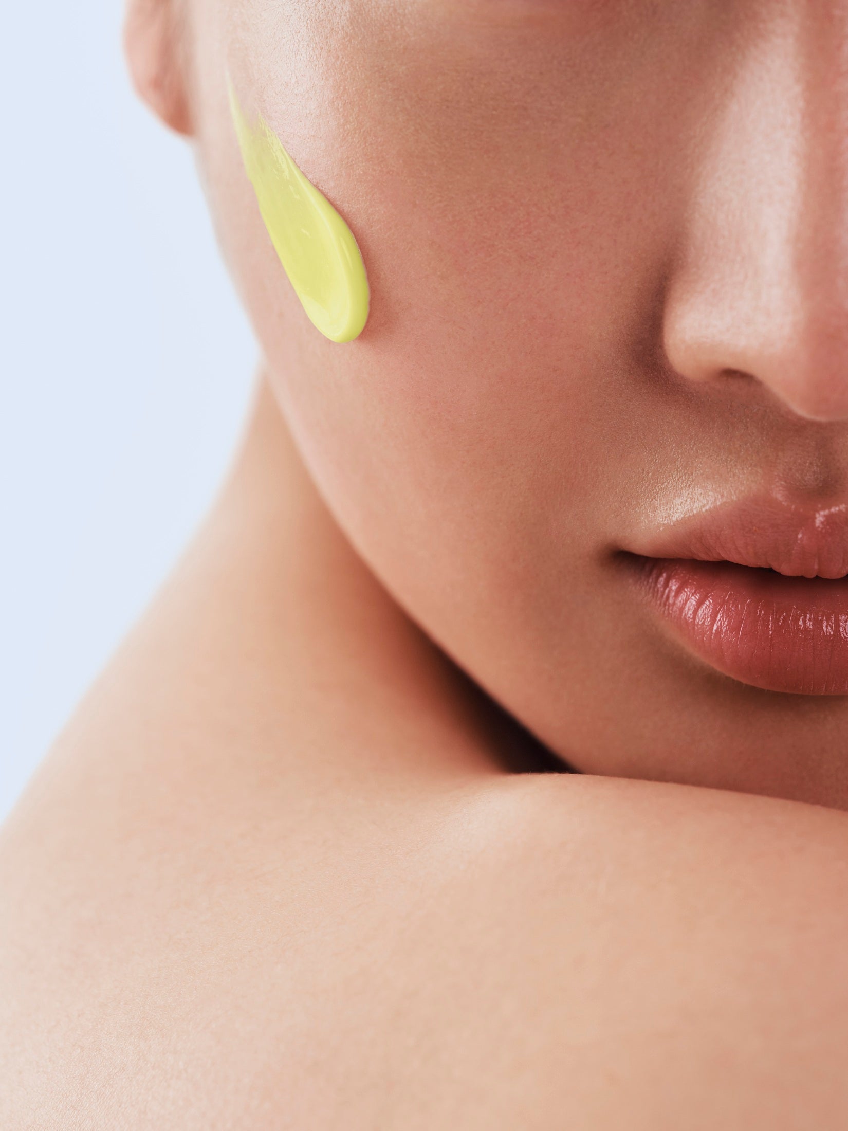 Close-up of a person's face with a green skincare product on their cheek against a light purple background