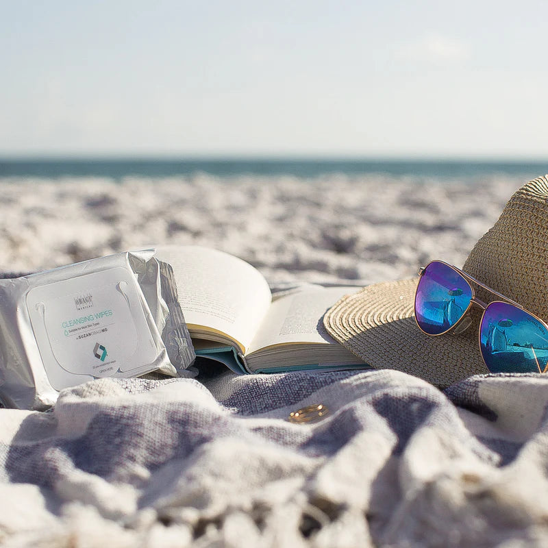 Open book, sunglasses, and hat on a beach with ocean view with Obagi cleansing wipes