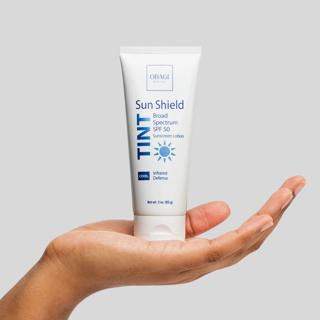 Obagi Sun Shield Tinted Sunscreen Lotion tube held in a hand against a gray background