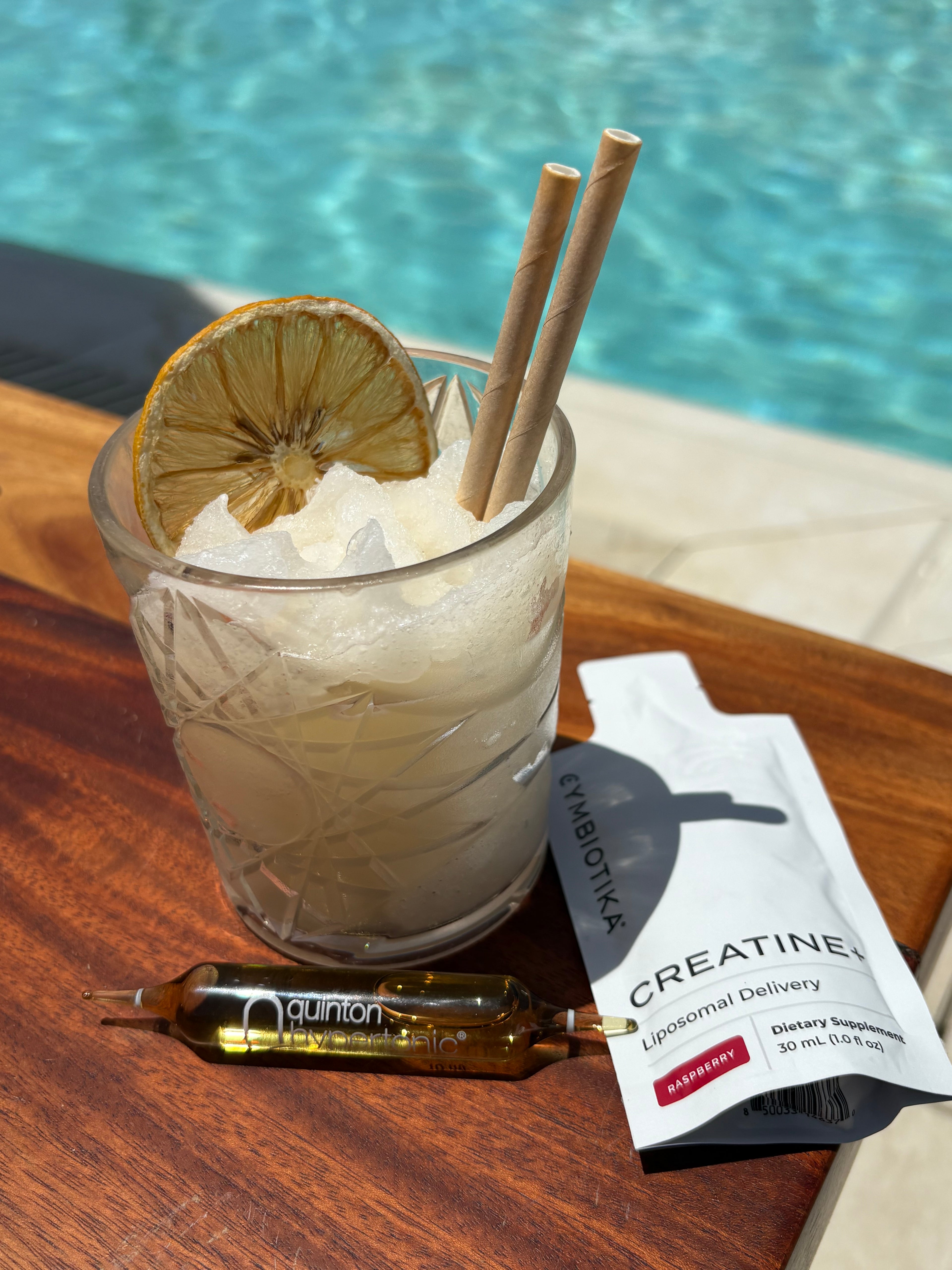 Glass with a cocktail, lemon slice, and two straws on a wooden table next to a bottle of Cymbiotika creatine.