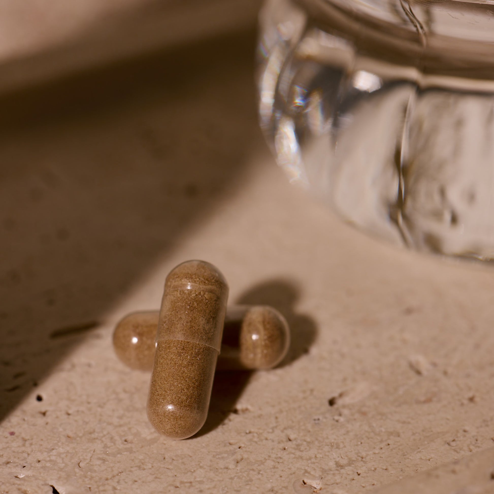 Three brown capsules on a sandy surface with a glass container in the background.